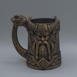 May include: Bronze-coloured Viking-themed beer mug. The mug features a detailed relief of a Viking warrior with a horned helmet and long beard. The handle is shaped like a dragon's head. The mug is designed with intricate patterns and details.