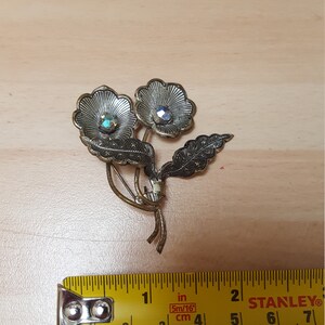 May include: A silver-toned brooch featuring two stylized flowers with iridescent rhinestones in the center. The flowers are connected by two leaves.