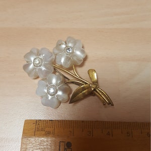 May include: A gold tone brooch with three white flowers and rhinestones. The flowers are made of a pearlescent material. The brooch is about 3 cm wide.