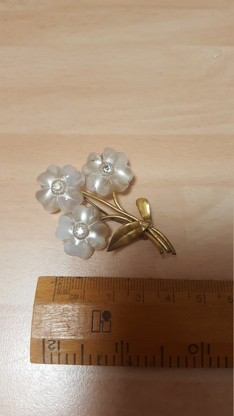 May include: A gold tone brooch with three white flowers and rhinestones. The flowers are made of a pearlescent material. The brooch is about 3 cm wide.