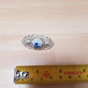 Può includere: Una spilla in filigrana d'argento con un cammeo in porcellana blu e bianca al centro. Il cammeo presenta un design stilizzato di un mulino a vento.