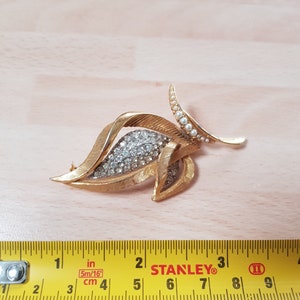 May include: A gold-toned brooch in the shape of a leaf with a cluster of clear rhinestones in the center. The leaf has a pearl-encrusted stem and is approximately 2 inches long.