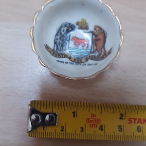 Op de afbeelding: Een klein wit keramiek schaaltje met een gouden rand en een wapenschild met een rode koe, een grijze hond en een bruine beer. Het wapenschild is omgeven door een blauw-wit lint met de tekst "Fortis Est Veritas" en "Arms of the City of Oxford".