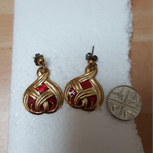 May include: A pair of gold-tone dangle earrings with red enamel accents. The earrings are shaped like a teardrop with a woven design.