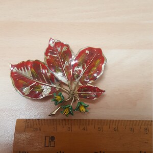 May include: A gold-tone brooch in the shape of three red leaves with green and yellow accents. The leaves have a metallic finish and are arranged in a cluster.