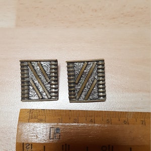May include: A pair of square-shaped earrings with a silver and gold finish. The earrings have a geometric design with a textured surface.
