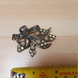 May include: A silver-toned brooch with a floral design. The brooch features a bow with a cluster of pearl-like beads and rhinestones.