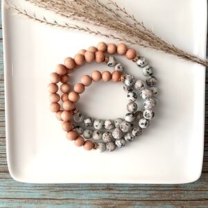 May include: Two stacked bracelets, one made of light brown wooden beads and the other made of white and grey speckled beads with gold accents.
