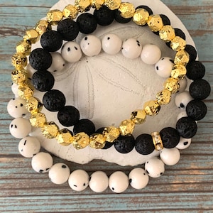 May include: Three beaded bracelets featuring black lava rock, white speckled, and gold-coloured beads. One bracelet has a small gold-coloured band with clear stones. The bracelets are displayed on a white, textured surface.