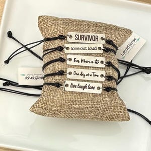 Peut inclure: Plusieurs bracelets avec des cordons noirs et des étiquettes métalliques rectangulaires. Chaque étiquette porte une phrase inspirante : "SURVIVOR", "love out loud", "Fur Mama", "One day at a Time" et "live laugh love". Les bracelets sont présentés sur un coussin en toile de jute.