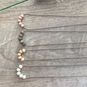 Peut inclure: Cinq colliers avec des perles en bois de différentes couleurs sur une chaîne argentée. Les perles sont disposées en forme de demi-cercle. Les colliers sont sur une surface en bois brun clair.