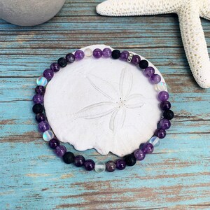 May include: A bracelet made of amethyst, black lava, and iridescent beads. The bracelet is circular and sits on a white sand dollar. The background is a blue and white wooden surface with a starfish and a grey pot.