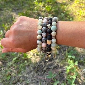May include: Beaded wrist keychains in shades of green, grey, black, and pink. Each keychain has a gold-coloured clasp. The bracelets are shown outdoors against a background of green foliage and grass.