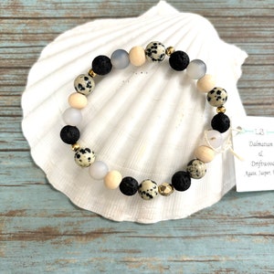 May include: A bracelet made of various natural stone beads, including black lava, speckled Dalmatian jasper, light wood, and frosted grey beads, with gold-coloured spacers. The bracelet is presented on a white seashell.