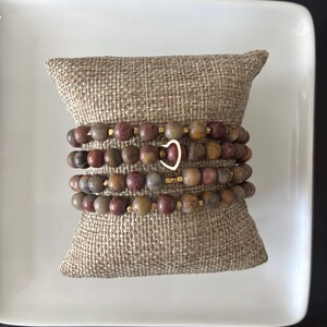 May include: Four beaded bracelets with earth-toned beads, displayed on a small burlap pillow. The bracelets feature a mix of brown, red, and tan beads, accented with gold-coloured spacers and a gold heart charm. The bracelets are presented on a white square plate.