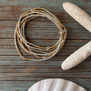 May include: A collection of gold and silver beaded bracelets arranged in a circular pattern. The bracelets sit on a distressed, blue-grey wooden surface. A white starfish and seashell are also present.