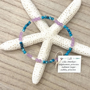 May include: A beaded bracelet featuring alternating round lilac amethyst and blue sediment jasper beads. A small white rectangular tag is attached with string, displaying the text: LS, Lilac Amethyst - enlightenment, protection, Sediment Jasper - stability, protection.