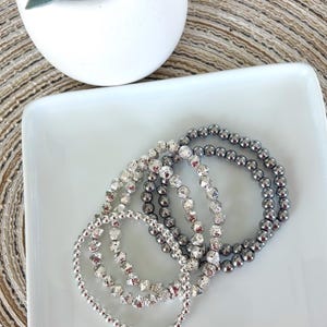 May include: A set of four elastic bracelets featuring round beads in varying textures and finishes. Two bracelets consist of smooth, polished dark grey beads, while the other two feature faceted, reflective silver beads that catch the light.