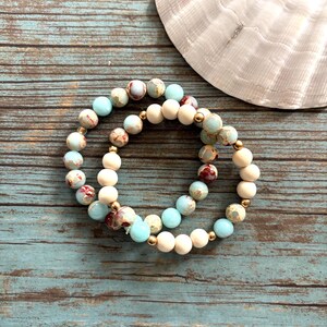 May include: Two beaded bracelets featuring light blue, cream, and patterned beads, interspersed with gold-coloured spacers. They rest on a weathered, turquoise wooden surface, with a white seashell in the background. The bracelets are a stylish accessory.