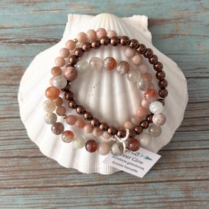 Peut inclure: Un ensemble de trois bracelets de perles composé de pierres de soleil rondes dans des tons pêche, beige et blanc translucide, alternées avec des perles en hématite bronze métallisé. Une étiquette blanche porte l'inscription Inner Glow, sunstone gemstones, bronze hematite.