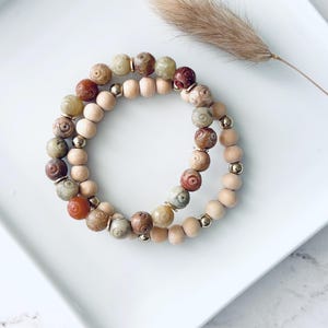 May include: Two bracelets with a natural, earthy aesthetic. One features light brown wooden beads, the other carved stone beads in brown, green, and red. Gold-coloured spacer beads add elegance. The bracelets are on a white surface.