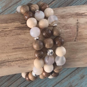 May include: A set of three bracelets featuring wooden beads in brown and beige tones, interspersed with white and light grey beads. The bracelets are stacked on a wooden surface.