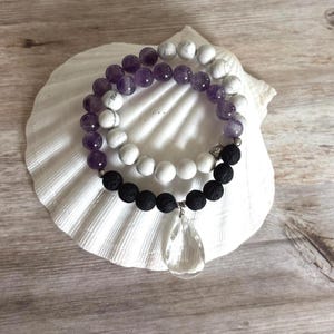 May include: A layered bracelet featuring a strand of purple amethyst beads and white marbled beads, paired with a strand of black porous lava beads. The bracelet includes a clear, teardrop-shaped crystal pendant and small silver-toned metal spacers.