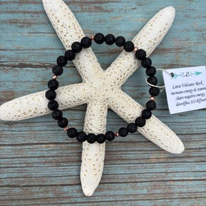 May include: A black lava rock bead bracelet with rose gold details, displayed on a white starfish against a blue wooden backdrop. The bracelet is a simple design. A tag reads "Lava Volcanic Rock".