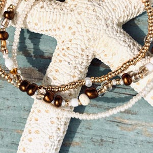 May include: Three beaded bracelets in gold, brown, and white rest on a white starfish against a weathered blue wooden surface. The bracelets feature small beads and a delicate design, perfect for beach or bohemian style.