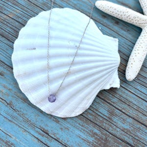 May include: A delicate silver necklace with a small, round, lavender gemstone pendant. The necklace is displayed on a large, white seashell against a backdrop of blue wooden planks, with a white starfish.