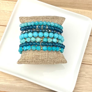 May include: A collection of blue and turquoise beaded bracelets. The bracelets are arranged on a small, burlap cushion, presented on a white rectangular plate and a wooden surface. The beads vary in shape and size, offering a diverse aesthetic.