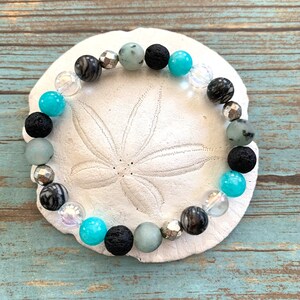 May include: A bracelet with a mix of stone beads in black, grey, turquoise, and clear. The bracelet includes faceted silver beads and is displayed on a white, textured surface. The beads are likely around 0.8 cm in diameter.