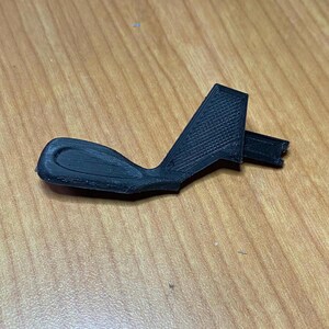 May include: A black plastic part with a textured surface and a curved handle. The part has a rectangular shape with a small, angled section on the top.
