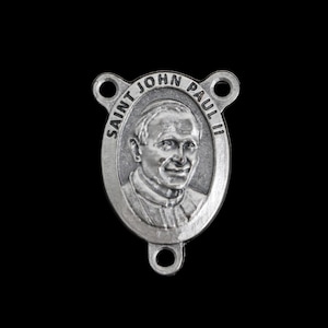 May include: A silver-colored religious pendant featuring an oval portrait of Saint John Paul II. The text "SAINT JOHN PAUL II" is inscribed around the portrait. The pendant has three holes for attachment and is set against a black background.