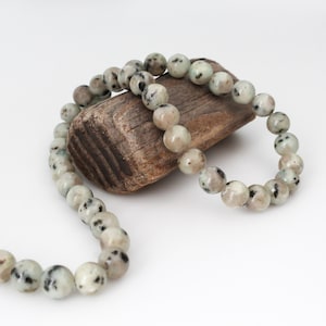 May include: A beaded necklace with speckled, light-colored stones. The necklace is draped over a piece of weathered wood against a white background. The beads are round and have a mix of gray, black, and cream colors.