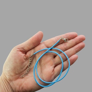 May include: A light blue necklace cord with a silver-colored clasp and chain extension. The cord is held in a hand against a gray background. The necklace cord is a simple design, suitable for pendants.