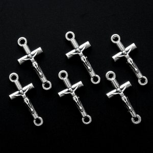 May include: Six silver-colored crucifix charms with a detailed depiction of Jesus on the cross. Each charm has a loop at the top and bottom for attaching to jewelry or other crafts. The charms are arranged on a black background.