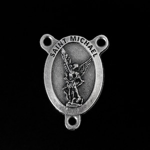 May include: A silver-colored Saint Michael rosary center with an oval medallion featuring an image of Saint Michael. The text "SAINT MICHAEL" is arched around the top of the oval. The piece has two holes for attachment.