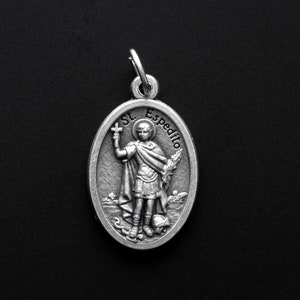 May include: Silver oval pendant featuring a depiction of Saint Expeditus, a Catholic saint, holding a cross and palm branch. The pendant is inscribed with "St. Expedito".