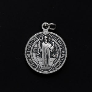 Puede incluir: Medalla de San Benito de plata con una imagen detallada del santo sosteniendo una cruz y un libro. La medalla tiene la inscripción "Eius in obitu nro. prae. sentia. muniamur" alrededor del borde.