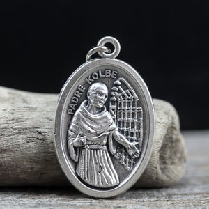 May include: Silver oval pendant with a depiction of Padre Kolbe, a Catholic saint, wearing a brown robe and holding a cross. The pendant features the words "Padre Kolbe" inscribed around the edge.