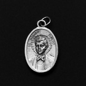 以下が含まれることがあります： 聖ドミニク・サヴィオの若いカトリックの聖人の詳細な画像が描かれた銀製のペンダント。ペンダントには、彼の頭上に後光が描かれ、「聖ドミニク・サヴィオ」という碑文が刻まれています。