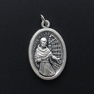 May include: Silver oval pendant with a depiction of Padre Kolbe, a Catholic saint, wearing a brown robe and holding a fence. The pendant has the words "Padre Kolbe" inscribed on it.