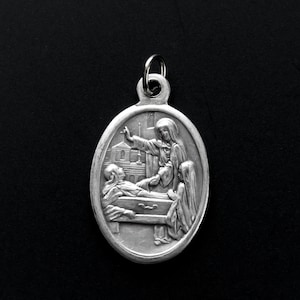 May include: Silver oval pendant with a religious image of a woman holding a cross and a man lying in a coffin. The pendant has a loop at the top for attaching to a chain.