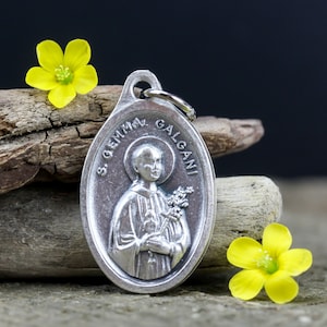 May include: Silver oval pendant featuring a depiction of Saint Gemma Galgani, a Catholic saint, holding a bouquet of flowers. The pendant is inscribed with the words "S. GEMMA GALGANI" around the edge.