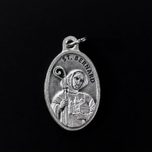 Könnte beinhalten: Silberner Anhänger mit einer Darstellung des Heiligen Bernhard, eines katholischen Heiligen, der einen Stab hält. Der Anhänger trägt die Inschrift "St. Bernard" am Rand.
