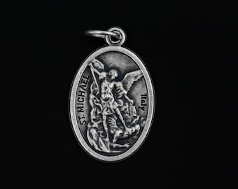 May include: Silver oval pendant featuring a detailed depiction of Saint Michael the Archangel slaying a demon. The pendant has the words "St. Michael" and "Italy" inscribed on it.