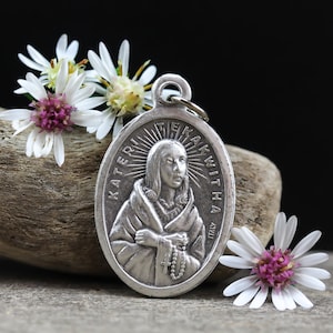 May include: Oval silver pendant featuring a detailed relief of a woman with the text "KATERI TEKAKWITHA" and "ITALY". The pendant includes a loop for a chain and a small rosary. White and purple flowers are arranged around the pendant.