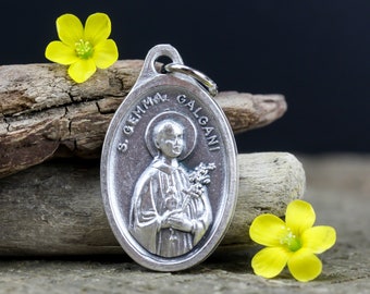 May include: Silver oval pendant with a depiction of Saint Gemma Galgani, a Catholic saint, holding a bouquet of flowers. The pendant has the words "S. GEMMA GALGANI" inscribed around the edge.