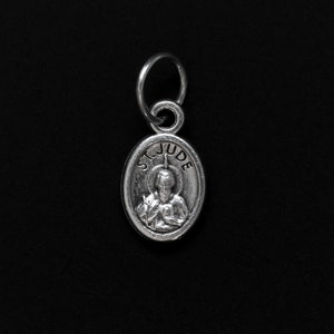 May include: Silver charm with a depiction of Saint Jude Thaddeus, patron saint of hopeless causes. The charm is oval shaped and has the words "St. Jude" inscribed around the image.
